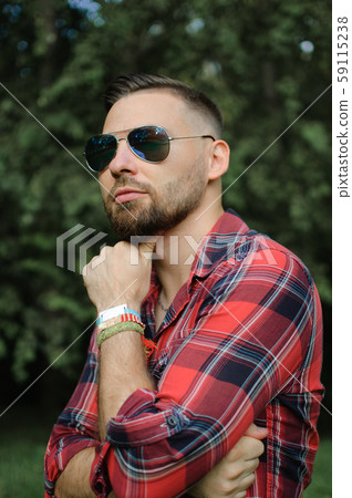 Outdoors portrait of young bearded man with sunglasses looking at the camera in the park on trees 59115238