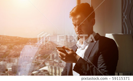 Young entrepreneur in office suit is browsing phone near the window with panoramic city view. 59115371