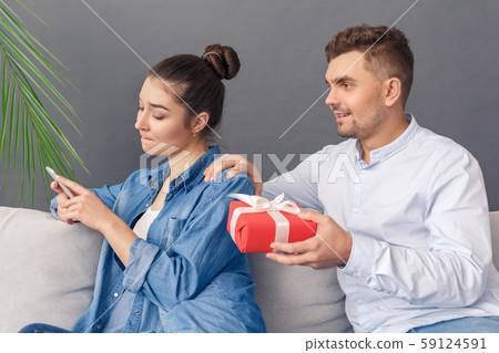 Modern Relationship Concept. Young couple man giving present to woman playing smartphone indifferent Modern Relationship Concept. Young couple man giving present to woman playing smartphone indifferent 59124591
