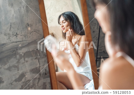 Weekend ar home. Young woman sitting on bed applying face powder smiling playful mirror reflection 59128053