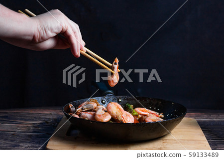 Chef at work. Hand holds a chopsticks under hot pan with fried vegetables, broccoli, cauliflower, carrots and shrimp. Process of preparation of vegetables over dark background Chef at work. Hand holds a chopsticks under hot pan with fried vegetables, broccoli, cauliflower, carrots and shrimp. Process of preparation of vegetables over dark background 59133489