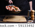 Chef at work. Hand holds a hot pan with fried shrimp and vegetables in sweet and sour sauce. Process of preparation of shrimps over dark background 59133492