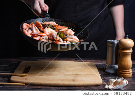 Chef at work. Hand holds a hot pan with fried shrimp and vegetables in sweet and sour sauce. Process of preparation of shrimps over dark background Chef at work. Hand holds a hot pan with fried shrimp and vegetables in sweet and sour sauce. Process of preparation of shrimps over dark background 59133492