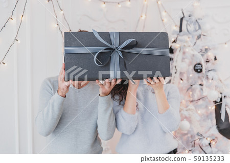 Shot of huge gray gift box with striped streamers. Blurred people hidding their faces behind the box Shot of huge gray gift box with striped streamers. Blurred people hidding their faces behind the box 59135233