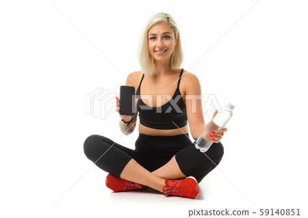 Beauiful sport caucasian girl sits on floor, cross legs, shows a mobile phone and keeps a bottle of 59140851