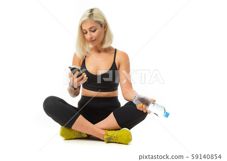 Beauiful sport caucasian girl sits on floor, cross legs, keeps a mobile phone and bottle of water Beauiful sport caucasian girl sits on floor, cross legs, keeps a mobile phone and bottle of water 59140854