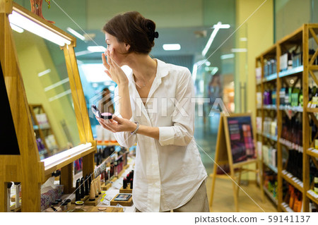 Young beautiful girl chooses her tone cream or powder and picks up on skin tone in front of big Young beautiful girl chooses her tone cream or powder and picks up on skin tone in front of big 59141137