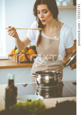 Young brunette woman cooking soup in kitchen. Housewife holding wooden spoon in her hand. Food and Young brunette woman cooking soup in kitchen. Housewife holding wooden spoon in her hand. Food and 59165141