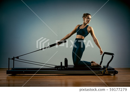 Young girl doing pilates exercises with a reformer bed. Beautiful slim fitness trainer on reformer Young girl doing pilates exercises with a reformer bed. Beautiful slim fitness trainer on reformer 59177279