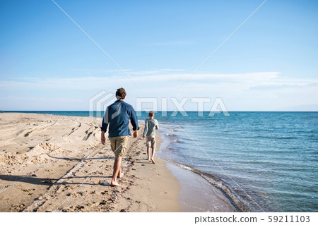 Rear view of father with small son outdoors on beach, running in water. Rear view of father with small son outdoors on beach, running in water. 59211103