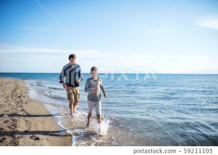 Father with small son on a walk outdoors on beach, running in water. 59211105