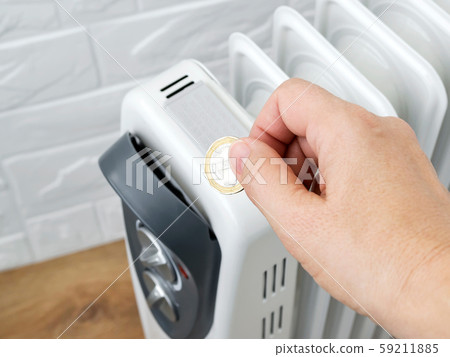 Woman hand putting a coin into home electric oil heater. The concept of higher electricity costs Woman hand putting a coin into home electric oil heater. The concept of higher electricity costs 59211885