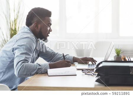 Black millennial employee using laptop in office and taking notes Black millennial employee using laptop in office and taking notes 59211891