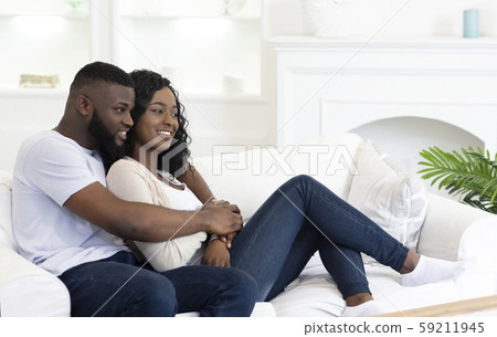 Happy black couple hugging while sitting on couch at home Happy black couple hugging while sitting on couch at home 59211945