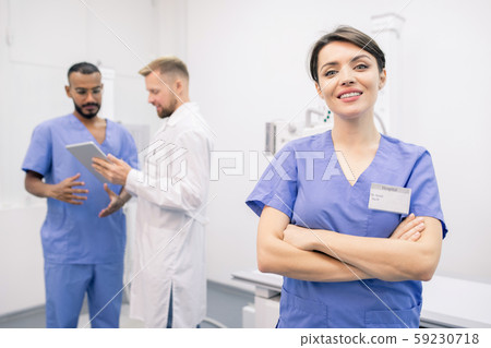 Happy young cross-armed clinician in uniform standing in front of camera Happy young cross-armed clinician in uniform standing in front of camera 59230718