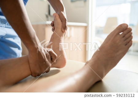 Hands of young mixed-race medical professional massaging one of feet of patient Hands of young mixed-race medical professional massaging one of feet of patient 59233112