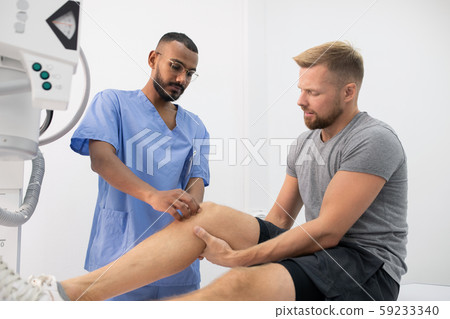 Young doctor in uniform massaging sick knee of one of patients in hospital Young doctor in uniform massaging sick knee of one of patients in hospital 59233340