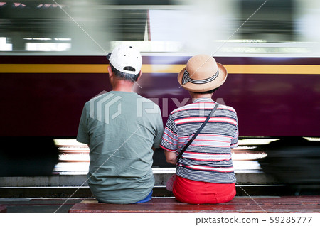 Back portrait of lovely adult couple sit on wooden bench and wait for the train approaching to the station with blurred motion of train in background Back portrait of lovely adult couple sit on wooden bench and wait for the train approaching to the station with blurred motion of train in background 59285777