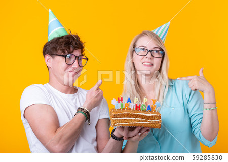 Young cheerful students charming girl and nice guy in greeting paper caps holding a cake. Concept of Young cheerful students charming girl and nice guy in greeting paper caps holding a cake. Concept of 59285830