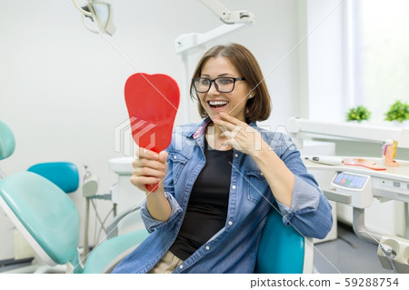 Happy woman patient looking in the mirror at the teeth, sitting in the dental chair 59288754