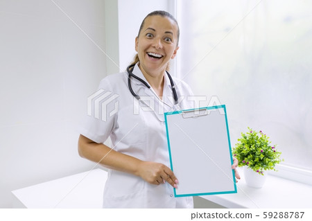 Adult smiling woman doctor with a blank sheet on clipboard for your text, background hospital window 59288787