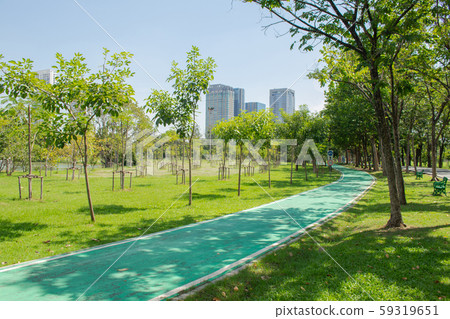 Green bicycle lane along side with green grass meadow field and trees in public park with city building in the background. Green bicycle lane along side with green grass meadow field and trees in public park with city building in the background. 59319651