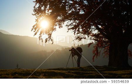 Hiker tourist man with camera on grassy valley on background of mountain landscape under big tree. Hiker tourist man with camera on grassy valley on background of mountain landscape under big tree. 59354639