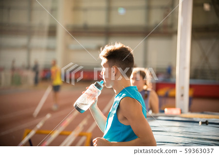 Young man in blue shirt standing on the sports stadium drinking a water from the bottle Young man in blue shirt standing on the sports stadium drinking a water from the bottle 59363037