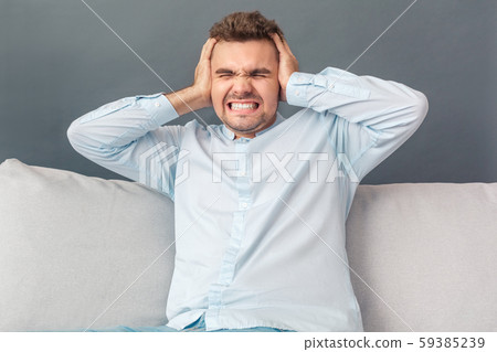 Freestyle. Young man studio sitting on sofa isolated on grey covering ears loud sound irritated 59385239