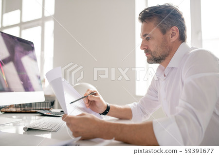 Mature serious engineer in white shirt pointing at sketch while checking it Mature serious engineer in white shirt pointing at sketch while checking it 59391677