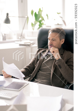 Serious director of company sitting by desk and looking through paper Serious director of company sitting by desk and looking through paper 59392023