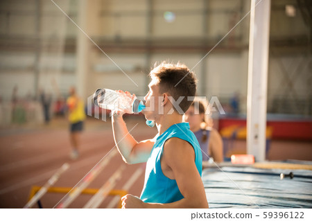 Young man in blue shirt standing on the indoors sports stadium drinking a water from the bottle Young man in blue shirt standing on the indoors sports stadium drinking a water from the bottle 59396122