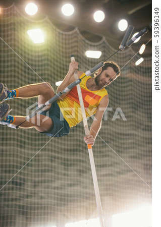 Pole vaulting - guy in yellow t shirt is jumping over the bar holding on to a pole Pole vaulting - guy in yellow t shirt is jumping over the bar holding on to a pole 59396149