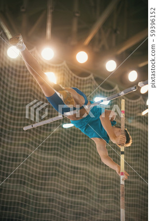 Pole vaulting - guy in blue t shirt is jumping over the bar holding on to a pole Pole vaulting - guy in blue t shirt is jumping over the bar holding on to a pole 59396152