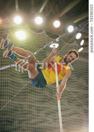 Pole vaulting in the sports stadium - an athletic smiling man in yellow shirt jumping over the bar Pole vaulting in the sports stadium - an athletic smiling man in yellow shirt jumping over the bar 59396503