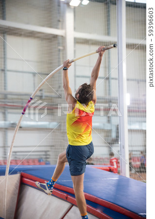 Pole vaulting indoors - a athletic man jumping over the bar - the pole flexing under the weight Pole vaulting indoors - a athletic man jumping over the bar - the pole flexing under the weight 59396603