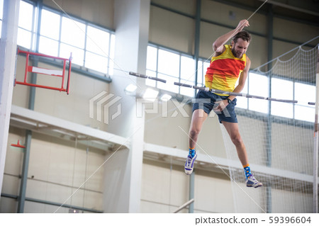 Pole vaulting indoors - a athletic man jumping over the bar and trying not to touch the bar Pole vaulting indoors - a athletic man jumping over the bar and trying not to touch the bar 59396604