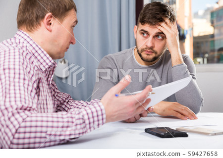Man worriedly discussing documents with friend at home table Man worriedly discussing documents with friend at home table 59427658