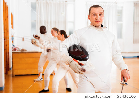 Active young male fencer in uniform standing with mask and foil at fencing room 59430100