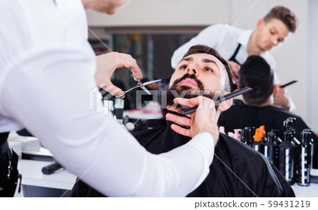 Male hairdresser accurately cutting beard of client 59431219