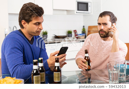 Two concerned men talking and laughing while enjoying beer at home, using phone Two concerned men talking and laughing while enjoying beer at home, using phone 59432013