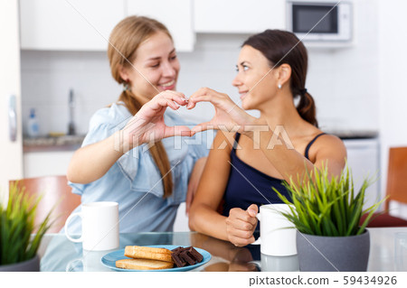 Young smiling emotional girlfriends showing the heart with hands and drinking tea Young smiling emotional girlfriends showing the heart with hands and drinking tea 59434926