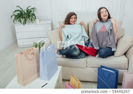 Young girls came back from the shop. They are sitting on the sofa, taking clothes out from the bags Young girls came back from the shop. They are sitting on the sofa, taking clothes out from the bags 59448267