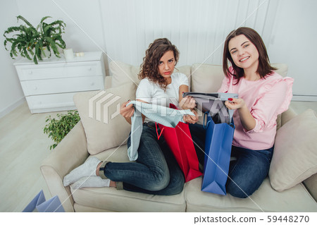 Cute schopahollic girls sitting on the sofa, taking out clothes from colorful shop bags. One girl is 59448270