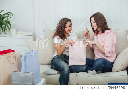 Two amazing happy friends sitting on the sofa, looking in one of the packets from the shop. A lot of 59448398