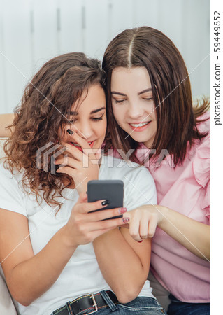 Beautiful young girls sitting on the sofa, at home, looking at the pictures in the phone, smiling 59449852