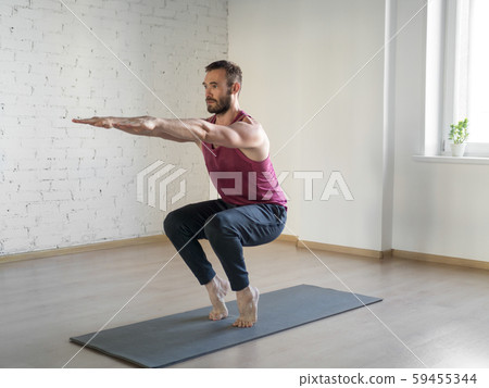 Chair pose. Caucasian fit man stands on tiptoe and doing yoga in fitness studio, selective focus. Chair pose. Caucasian fit man stands on tiptoe and doing yoga in fitness studio, selective focus. 59455344