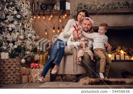 Photo of happy parents in Santa's cap with sons at fireplace near tree with garland in room 59456636