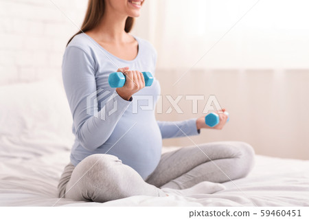 Unrecognizable Pregnant Lady Exercising With Dumbbells Sitting On Bed, Cropped Unrecognizable Pregnant Lady Exercising With Dumbbells Sitting On Bed, Cropped 59460451