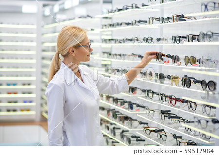 Blonde eye doctor putting different glasses on shelves in store 59512984